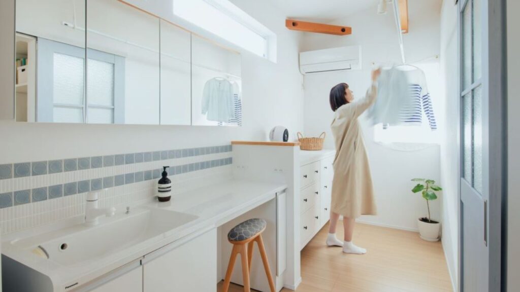 くらはし建築の注文住宅に設置された、脱衣所兼ランドリールームの施工事例。白い壁と木のカウンターでデザインされたおしゃれな空間で、女性が洗濯物を部屋干ししている。天井にはエアコンと物干し竿が設置されており、梅雨や花粉の季節でも洗濯物がすぐ乾く、家事ラクな暮らしを実現している。