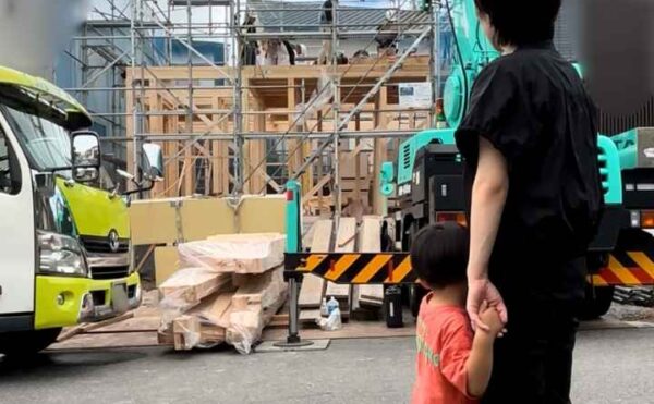 我が家が建つ上棟の様子を、感慨深く見つめるお施主様（お母さん）と、その手をしっかり握る小さなお子さんの後ろ姿。