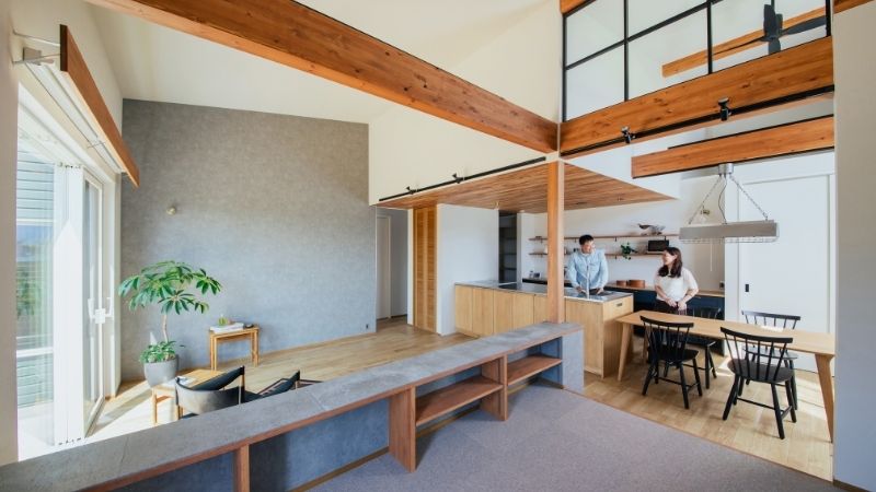 くらはし建築の施工事例写真。勾配天井と木の梁が印象的な、開放感あふれる平屋のLDK。大きな窓から光が差し込む心地よい空間で、ご夫婦がキッチンに立つ、豊かな暮らしのワンシーンです。