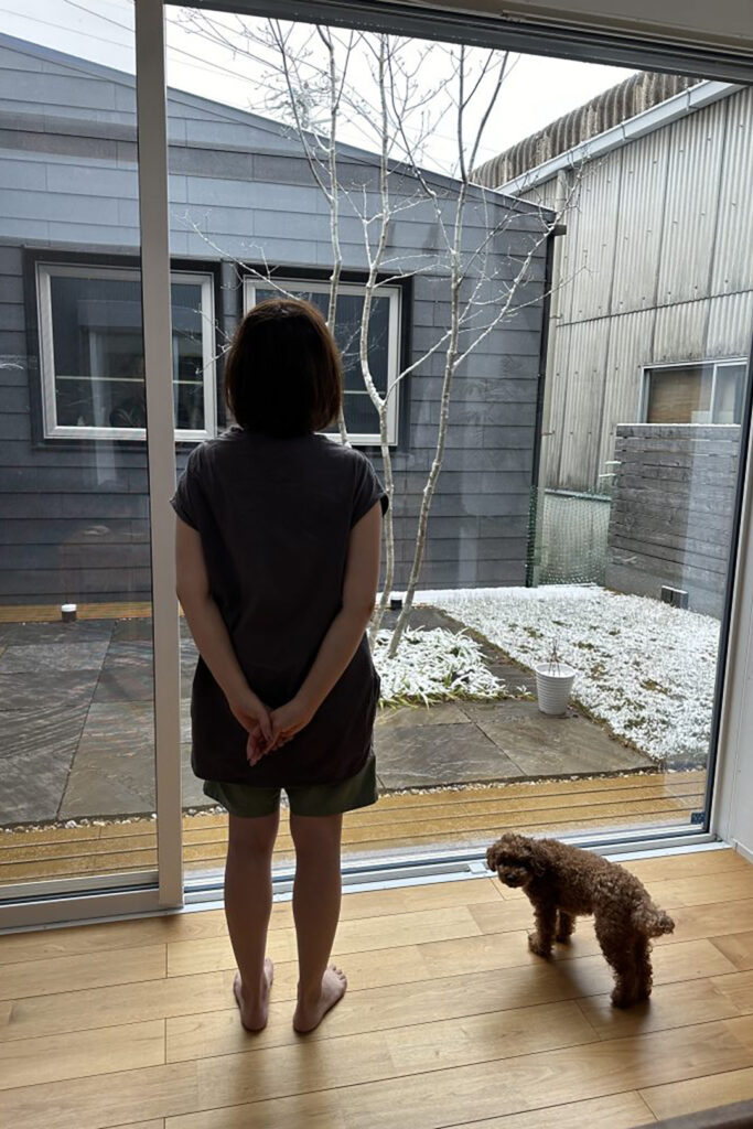 雪が積もる庭を暖かい室内から半袖・裸足で眺める女性。くらはし建築の家が実現する、冬でも春のように快適な暮らし。