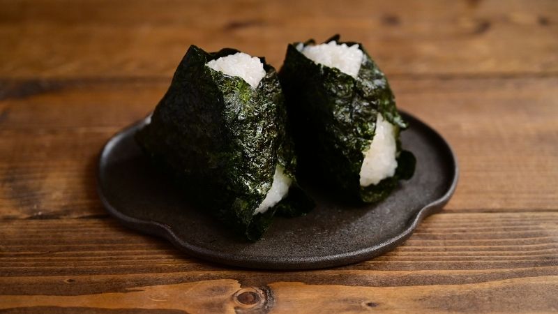 木目調のテーブルに置かれた、海苔が巻かれた2つの美味しそうなおにぎり。IHクッキングヒーターでは直火がないため、海苔を炙ることができないという「IH派あるある」の悩みを象徴する一枚。